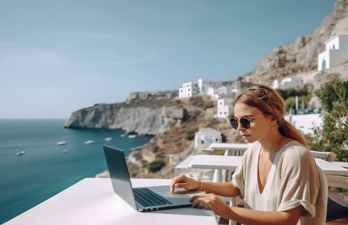 A lady with a loose ponytail and sunglasses sitting outside a cafe at her laptop with a view of the ocean.
