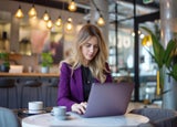 A blonde woman with a purple blazer sitting at a cafe table working on a laptop.