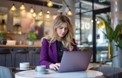 A blonde woman with a purple blazer sitting at a cafe table working on a laptop.