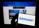 A pair of hands holding a smartphone device displaying the Ticketmaster logo with a backdrop of a computer screen of the company website.