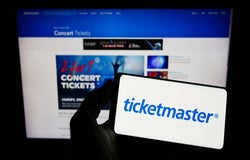 A pair of hands holding a smartphone device displaying the Ticketmaster logo with a backdrop of a computer screen of the company website.