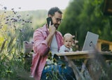 Man working on a laptop and talking on a phone while holding a baby