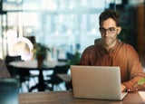 A man in a brown shirt with glasses and dark brown hair sitting at an office desk with his laptop.