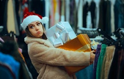 A woman with a light brown fur coat and red Santa hat carrying two large Christmas presents in the middle of a clothing store.