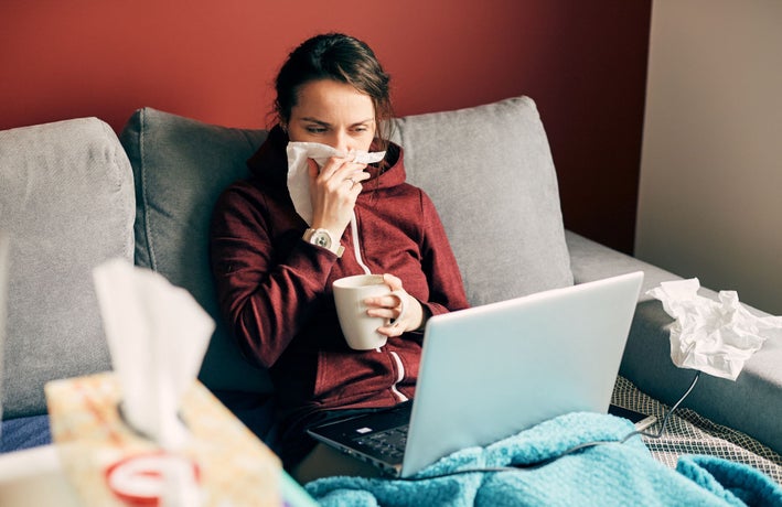 A woman with a burgundy hoodie holding a cup of tea and a tissue to her nose. She is sitting on a grey sofa with a grey laptop on her lap.