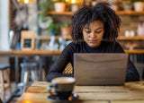 A woman with a black sweater sitting at wooden cafe table with a brown laptop.