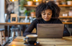 A woman with a black sweater sitting at wooden cafe table with a brown laptop.