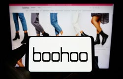 A smartphone device displaying the Boohoo logo against a backdrop of a computer screen with Boohoo product listings.