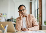 A man with black hair, black rimmed glasses wearing a plain white t shirt and a light peach jacket sitting at a work desk looking bored.