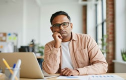 A man with black hair, black rimmed glasses wearing a plain white t shirt and a light peach jacket sitting at a work desk looking bored.