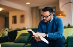A man with a blue sweater sitting on the coach reading a book.