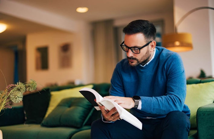A man with a blue sweater sitting on the coach reading a book.