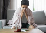 A man sitting on a grey sofa blowing his nose with a tissue