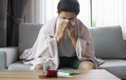 A man sitting on a grey sofa blowing his nose with a tissue
