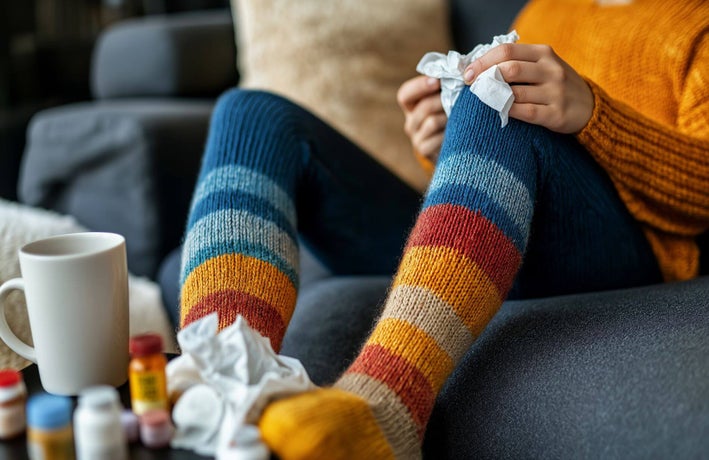 A shot of someone sitting on the sofa resting their feet on a coffee table filled with medicine and tissues.