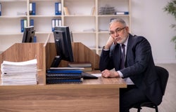 A man in a black suit sitting at a wooden office desk looking bored.