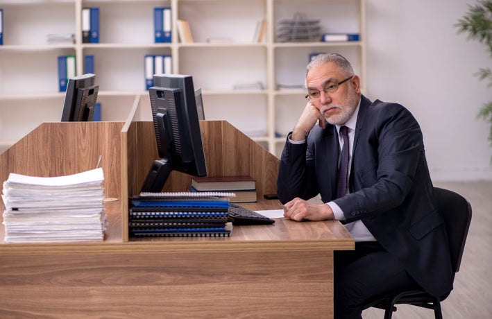 A man in a black suit sitting at a wooden office desk looking bored.