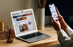 A person uses their smartphone to check something, while their laptop sits open on their desk