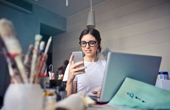 A young woman in a work studio is using her smartphone in front of a laptop